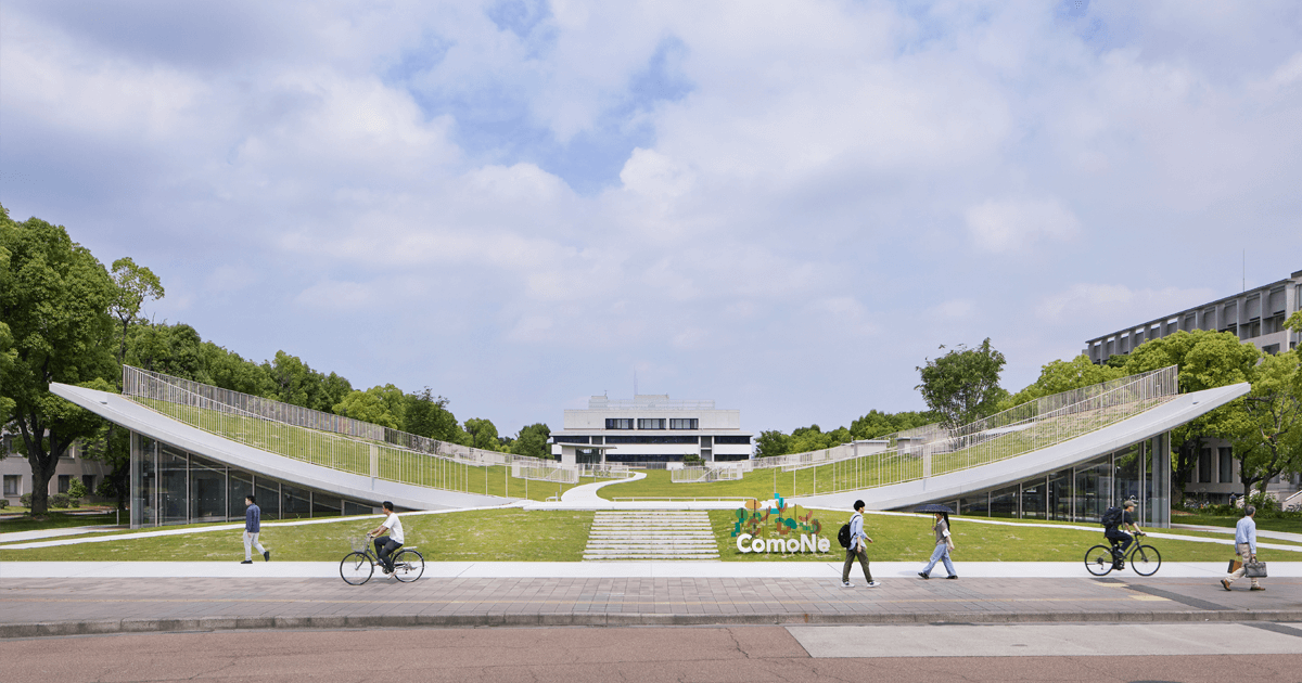 画像：都市型開放的なスペースで緑豊かなエリアと現代的な建物が特徴的なComoNe施設の外観。歩道を歩く人々やバス、車が行き交う風景。