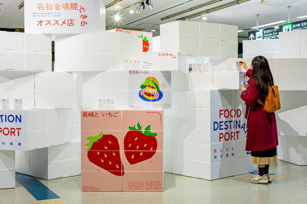 長崎空港内で開催された「FOOD DESTINATION PORT」の展示風景。白い箱を積み上げて構成されたブースに、長崎の食文化を紹介するグラフィックが貼られている。中央には赤いイチゴのイラストと「長崎といちご」の文字があり、右側では赤いコートを着た来場者が展示の一部を手に取って見ている。上部には「名物を堪能できるオススメ店」などのキャッチコピーが掲げられている。