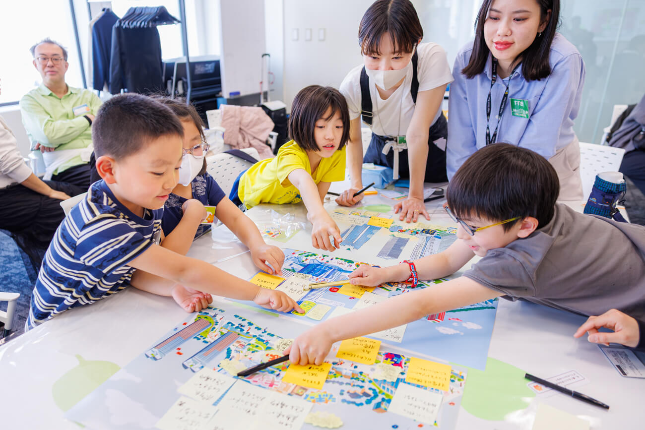 子どもたちが机を囲み、ワークショップを行っている様子。メモを指差しながら議論をしている