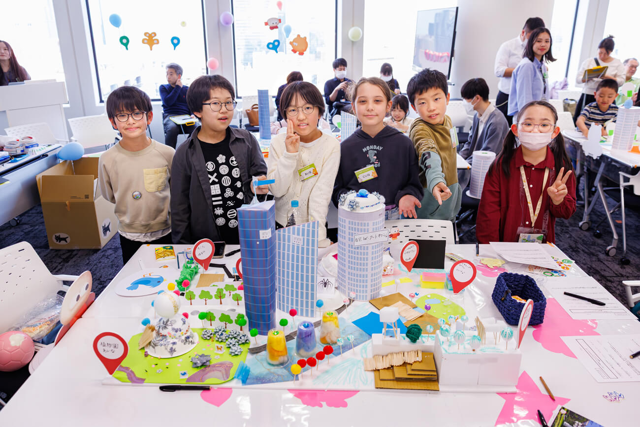 完成したまちの模型の前で写真を撮る、ワークショップに参加した子どもたちの写真。まちの模型の中にはビルと、植物園や公園が配置されている