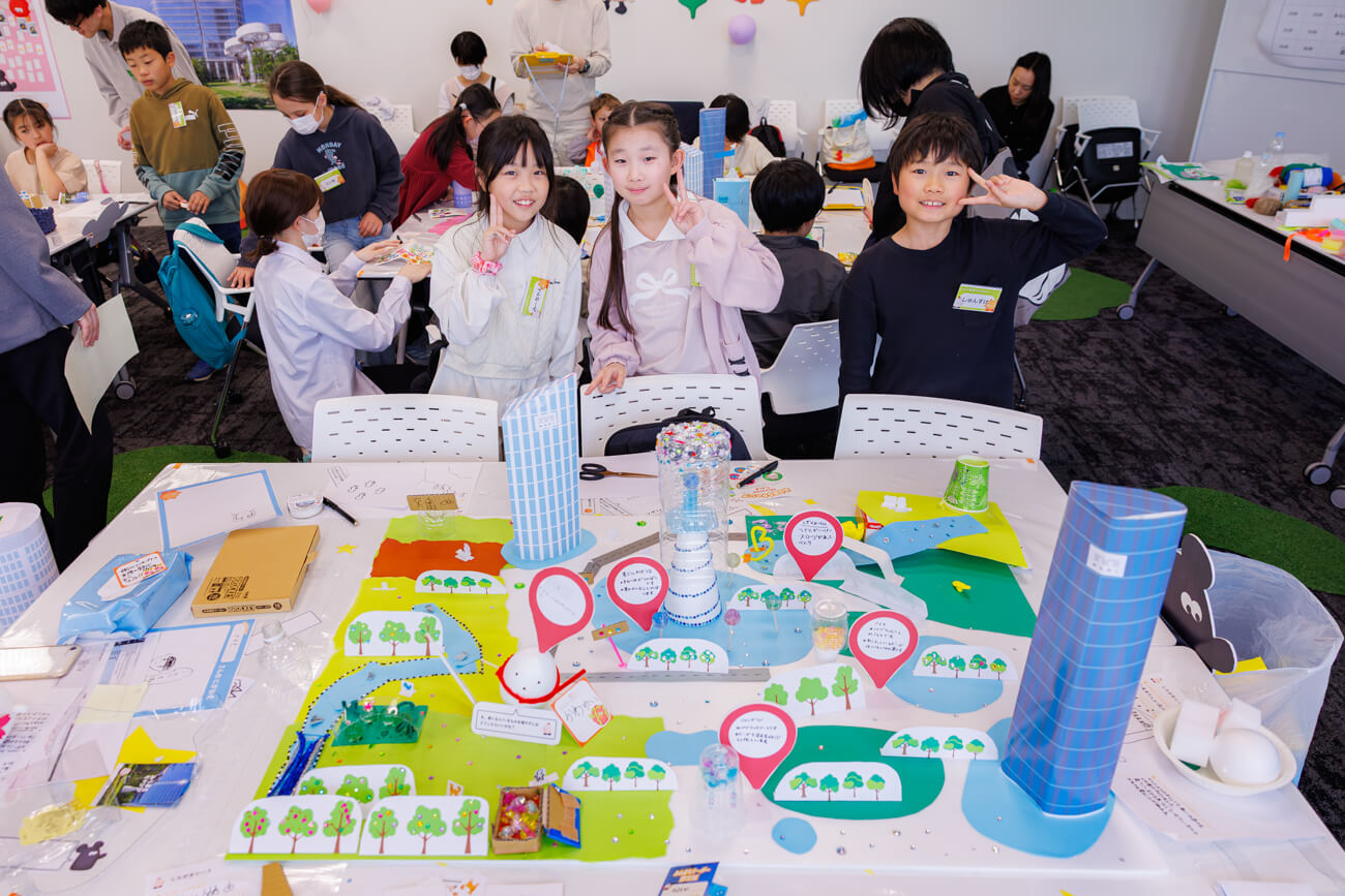 完成したまちの模型の前で写真を撮る、ワークショップに参加した子どもたちの写真。まちの模型の中には緑の広場やたくさんの木々、噴水などが配置されている