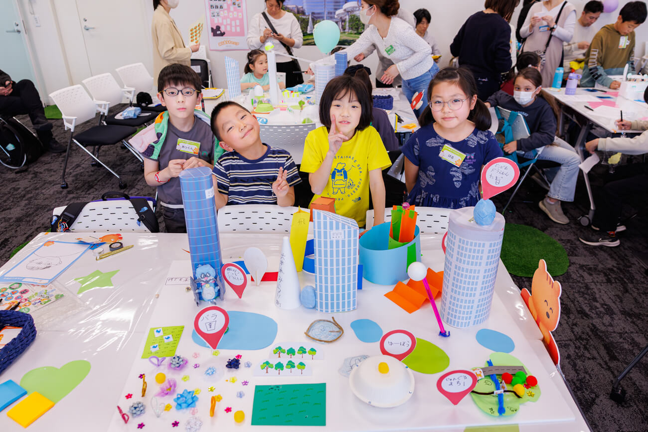 完成したまちの模型の前で写真を撮る、ワークショップに参加した子どもたちの写真。まちの模型の中にはバラ園のほか、「ゆーほう（UFO）」と書かれた造形物が