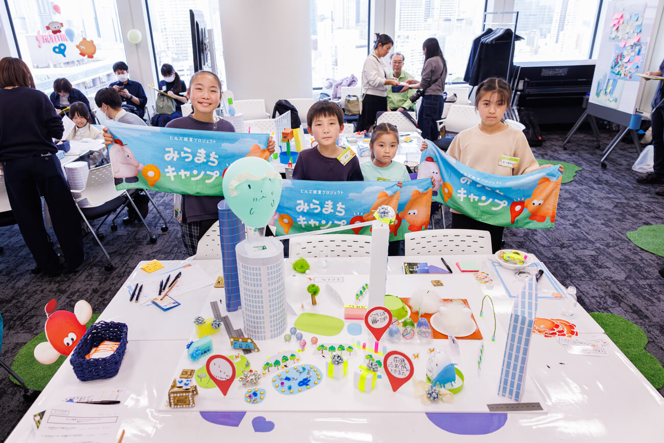 完成したまちの模型の前で写真を撮る、ワークショップに参加した子どもたちの写真。まちの模型の中にはビルや木々が並び、「ぷれぜんとのまち」と書かれたメモが見える