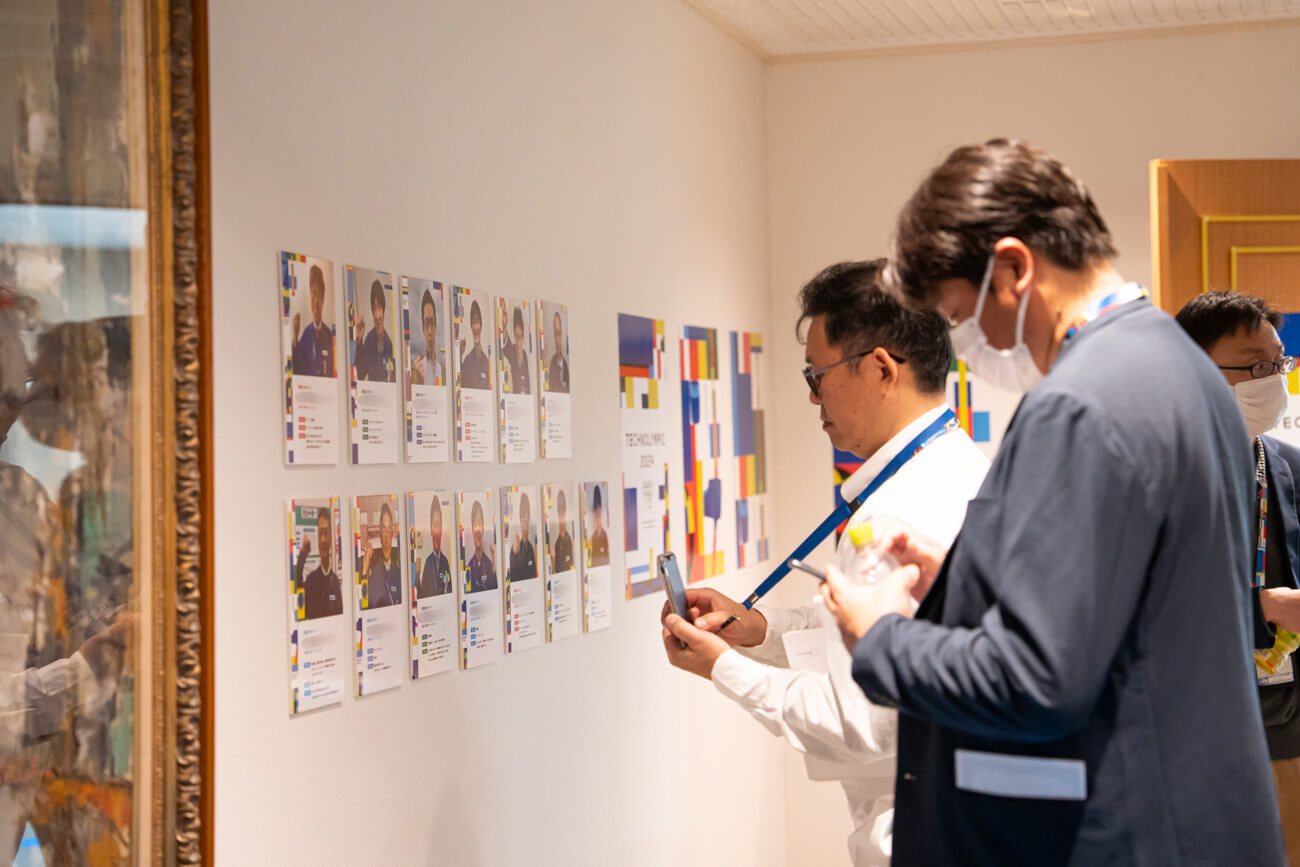 壁に掲示された出場選手のプロフィールを眺める、スーツス姿の男性たちの写真