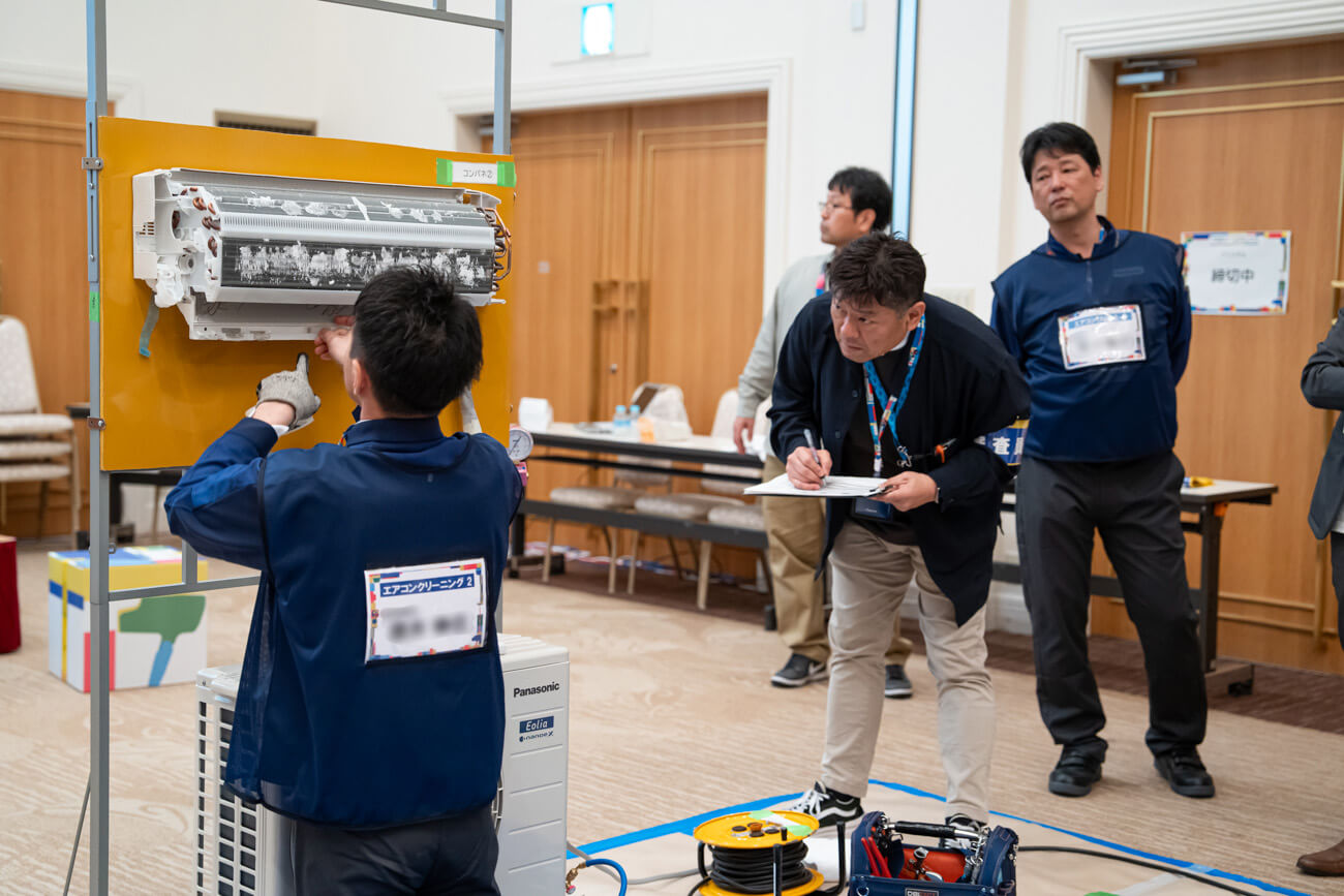 家電を修理する技術員と、その様子を見守りながら手元の採点ボードらしきものに何かを記入するスタッフの写真