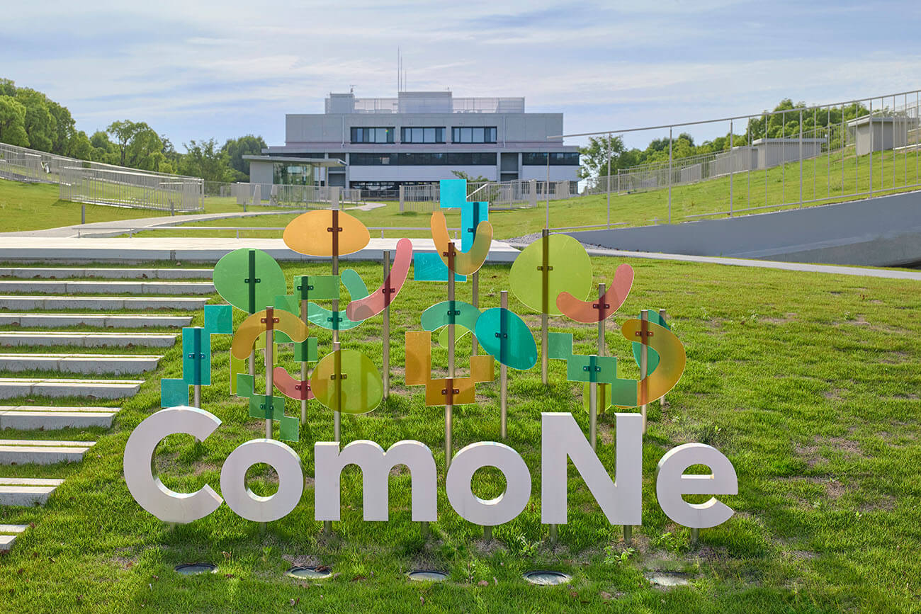 芝生広場に設置された屋外サイン。色とりどりの透明なパネルと立体文字が組み合わさり、背景には建物が見えている。