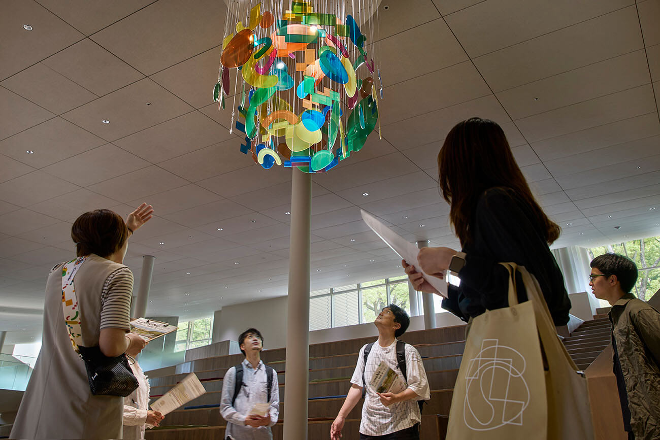 館内ツアーで、クルーが天井から吊るされたカラフルなインスタレーションを指し示しながら来館者に解説している様子。参加者は手元の資料を見たり、作品を見上げたりしながら説明に耳を傾けている。