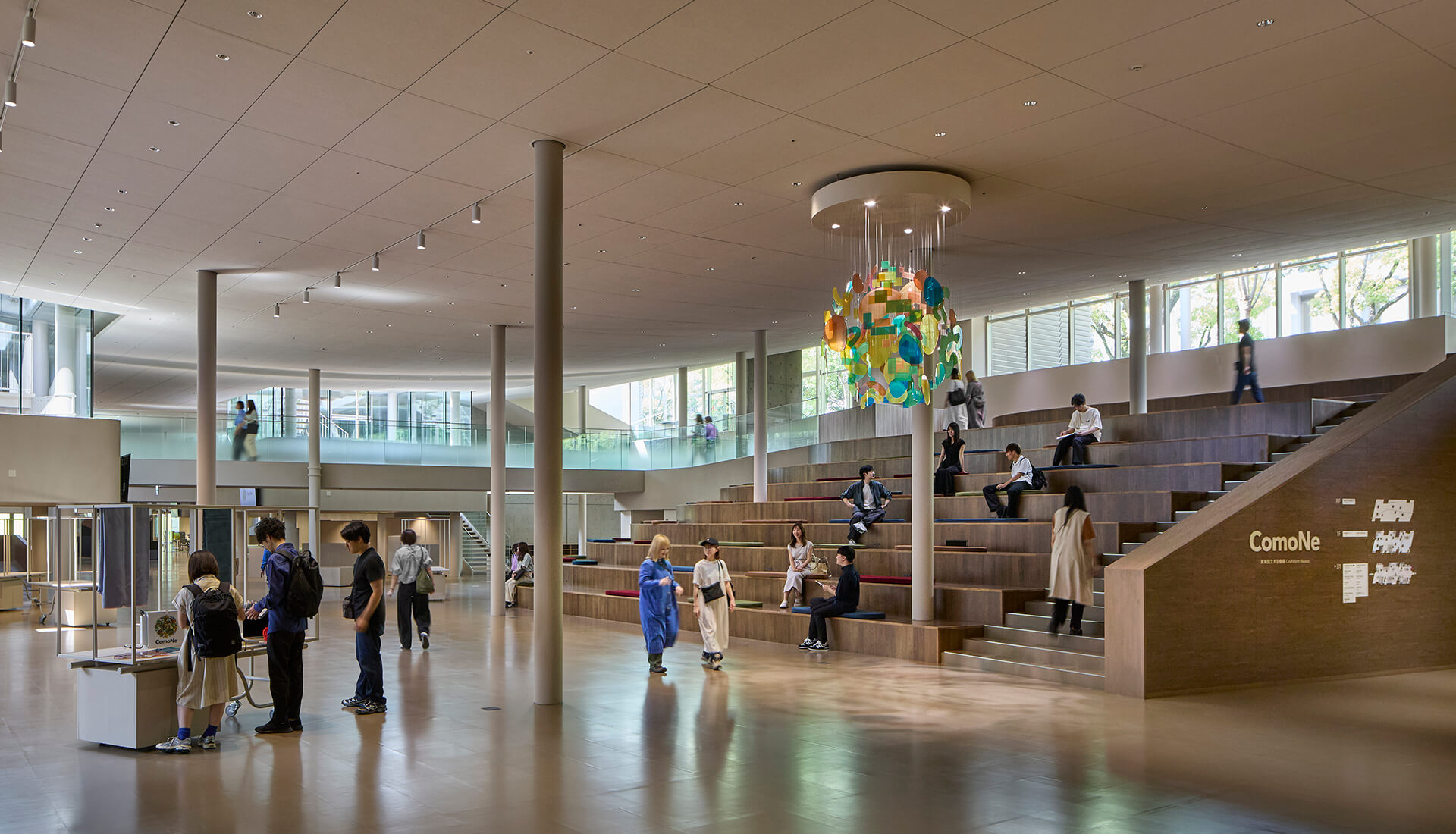 『ComoNe』内観。広々とした吹き抜け空間に大階段状の観覧席が設けられ、学生や来館者が思い思いに過ごしている。中央には色鮮やかなオブジェが吊り下げられている。