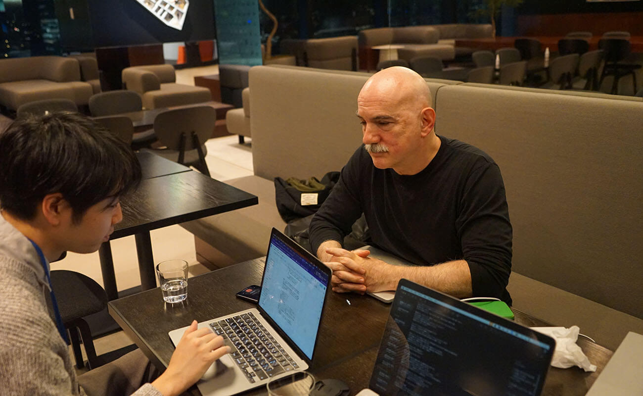 KMDの教員にインタビューを行う様子。教員はテーブル越しに座り、インタビュアーの話に真剣に耳を傾けている。テーブル上にはノートパソコンとグラスが置かれており、リサーチ活動の一環としての対話の場面が切り取られている。