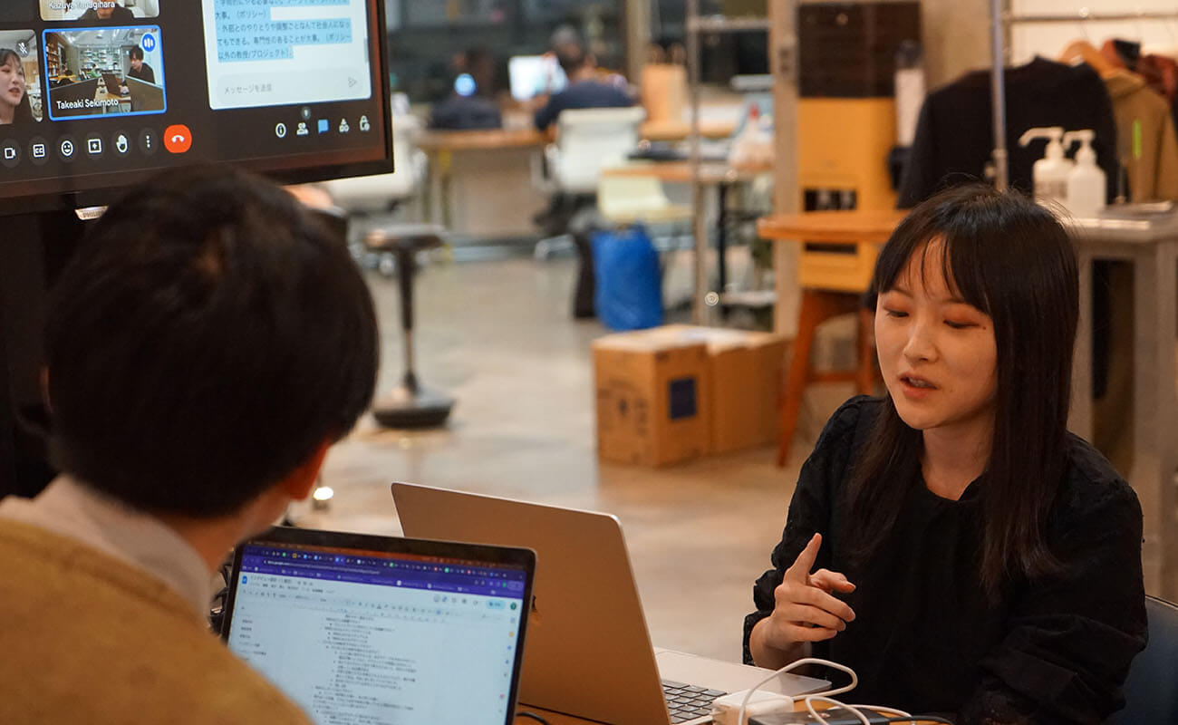 KMDの卒業生に対するインタビューの様子。卒業生が身振りを交えながら語る姿が映されており、ノートパソコンを前に聞き手が話を記録している。モニターにはオンライン参加者の画面も表示されており、対面とオンラインを組み合わせたハイブリッド形式のリサーチが行われていることがわかる。