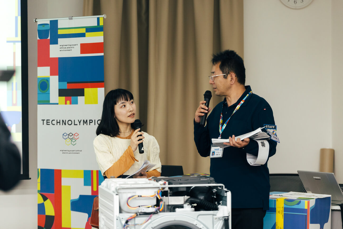 競技会で司会をする男女の写真。二人の背後には、キービジュアルをモチーフとした会場装飾がある