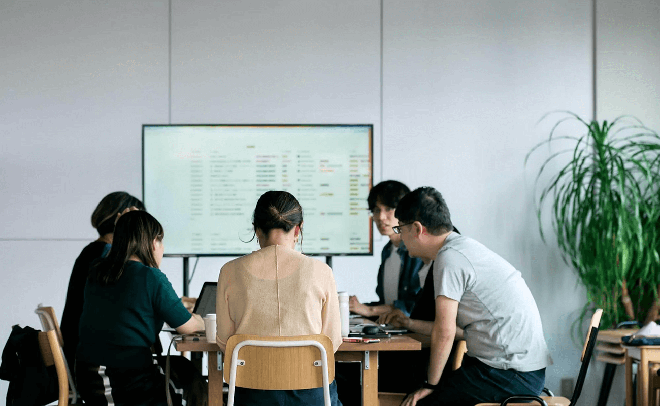 会議室で5人のメンバーが机を囲み、ノートPCを開きながら議論している。背後のモニターには検討項目のリストが映し出されている。