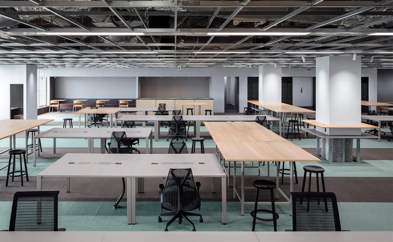 新オフィス執務エリア。白と木目の机やスツールが並び、開放的でフレキシブルな働き方に対応する空間が広がっている。