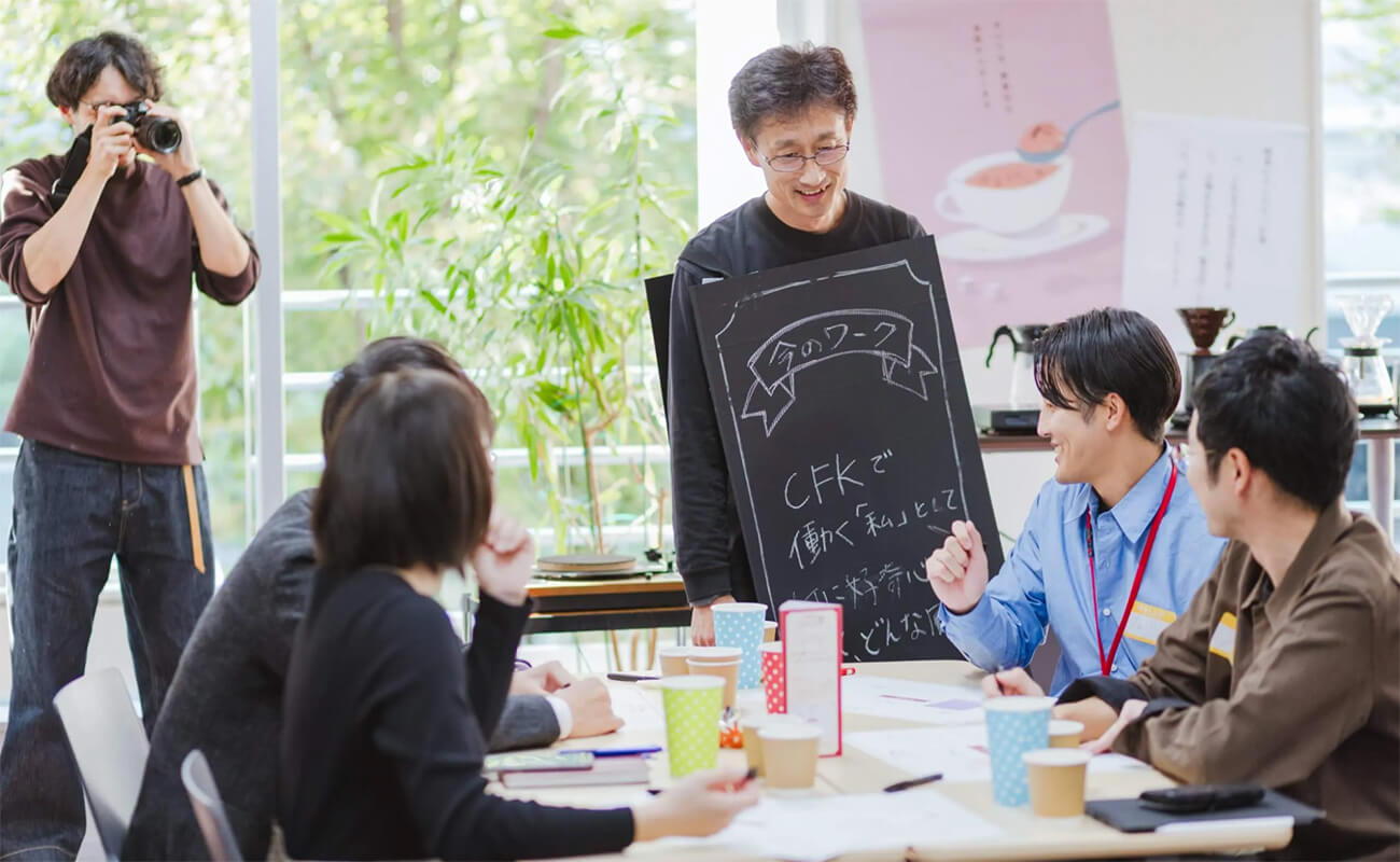 CFKのワークショップ風景。社員が黒板に書いたメッセージを示しながら発表し、周囲の参加者が笑顔で意見を交わしている。