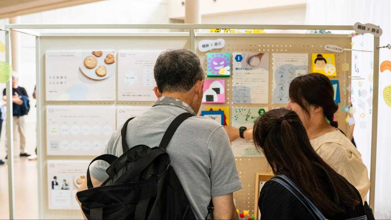 大きなパネルの前にプロジェクトの担当者が立ち、二人の来場者に対して説明をしている様子