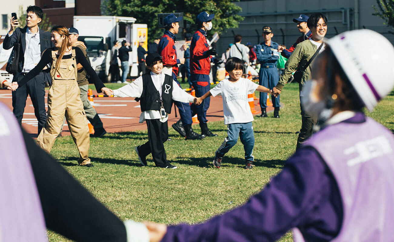 屋外の防災訓練プログラムで、大人と子どもたちが手をつなぎ円になって活動している様子