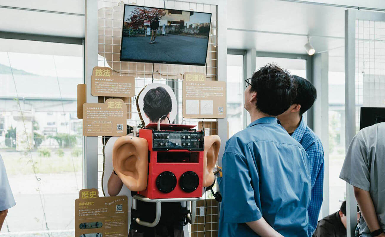展示会場で、大きな耳の模型を取り付けた赤い装置の展示を来場者が覗き込み、上部のモニター映像を見ている