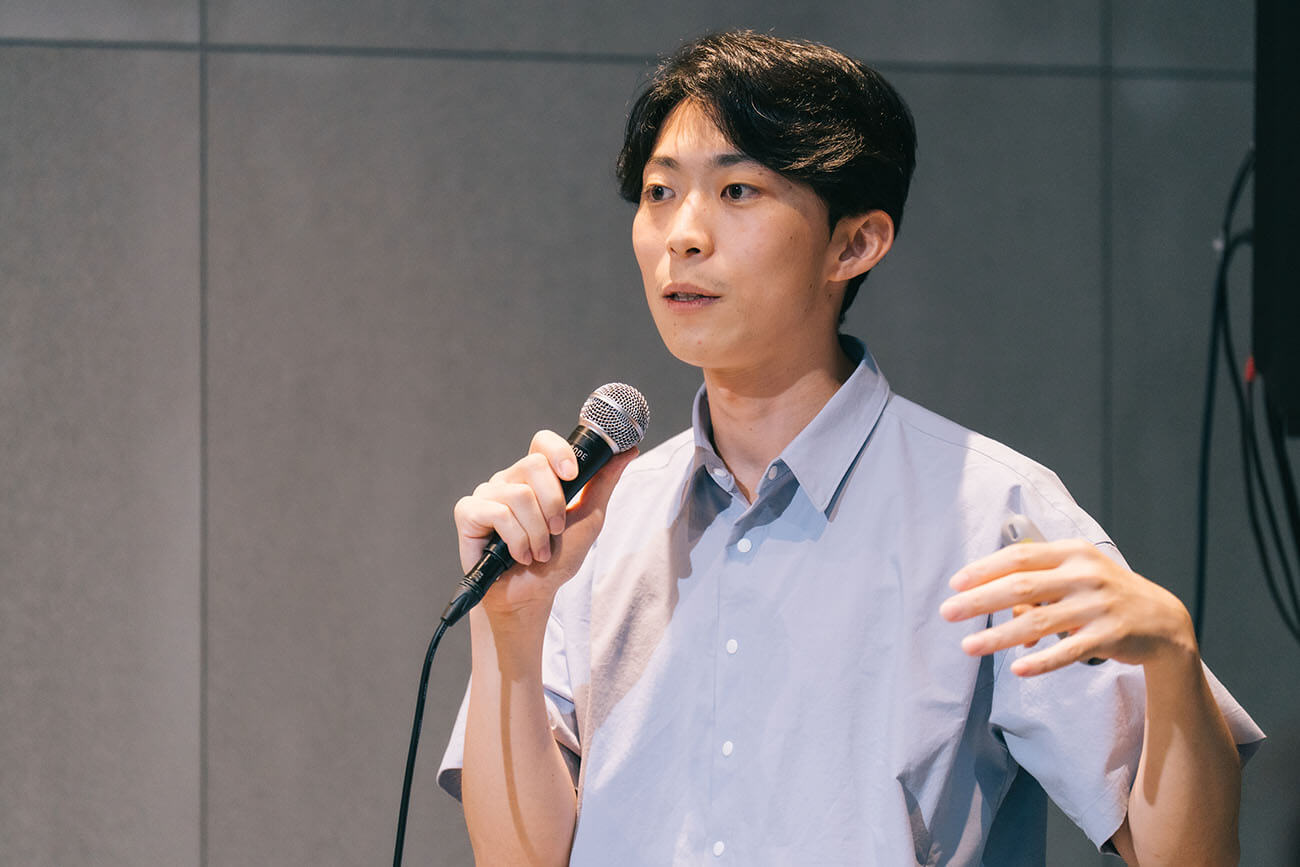 マイクを持ち、堂々と登壇する男性のバストアップ写真（中圓尾）