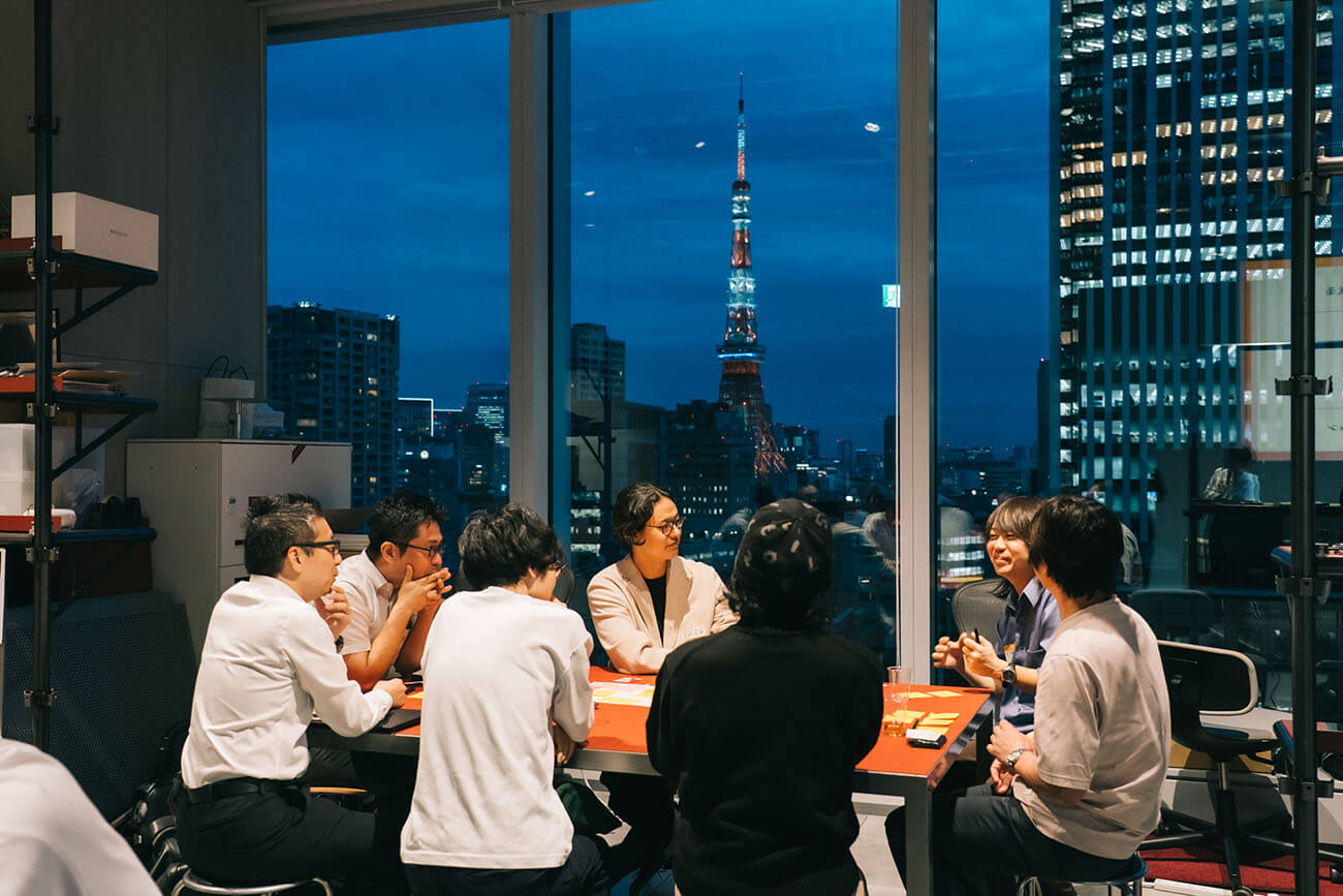 オープンディスカッションでの、ひとつのテーブルでの一幕を捉えた写真。男性参加者の話をグループ一同で聞きいいっている様子。背景には、夜になった都会の風景に東京タワーが聳え立っている。