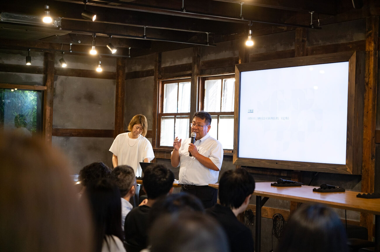 白シャツ姿の男性がマイクを持ち話している様子