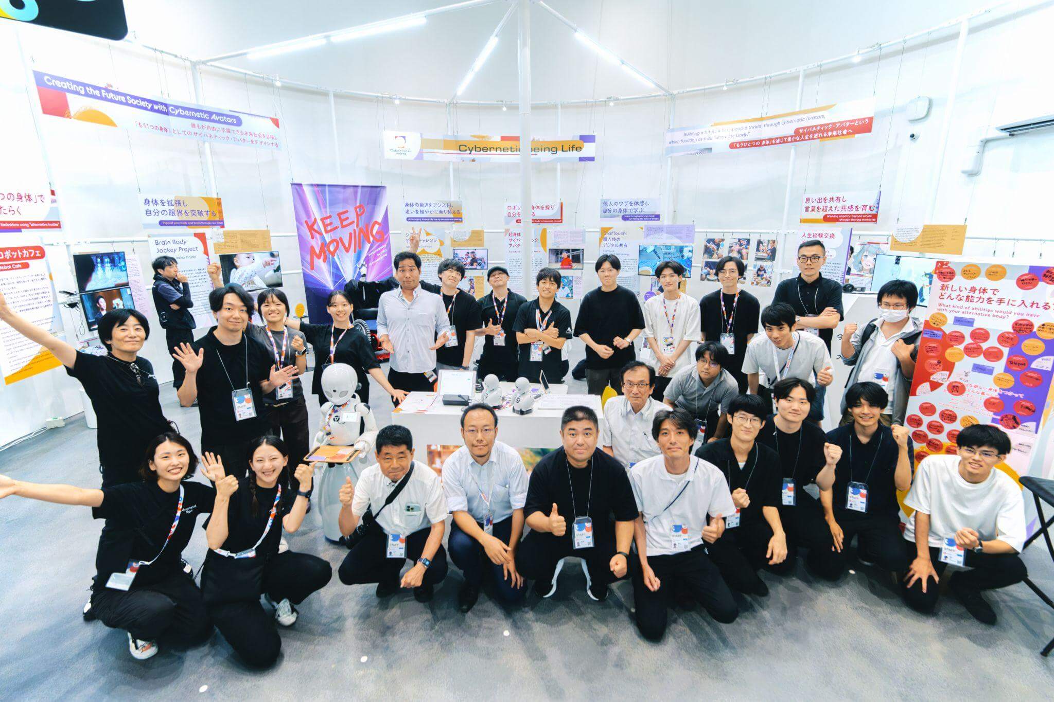 展示会場でスタッフや研究者たちが一堂に並び、笑顔でポーズを取る集合写真。体験展示を支えた多様なメンバーがそろい、達成感を味わっている雰囲気が伝わる。