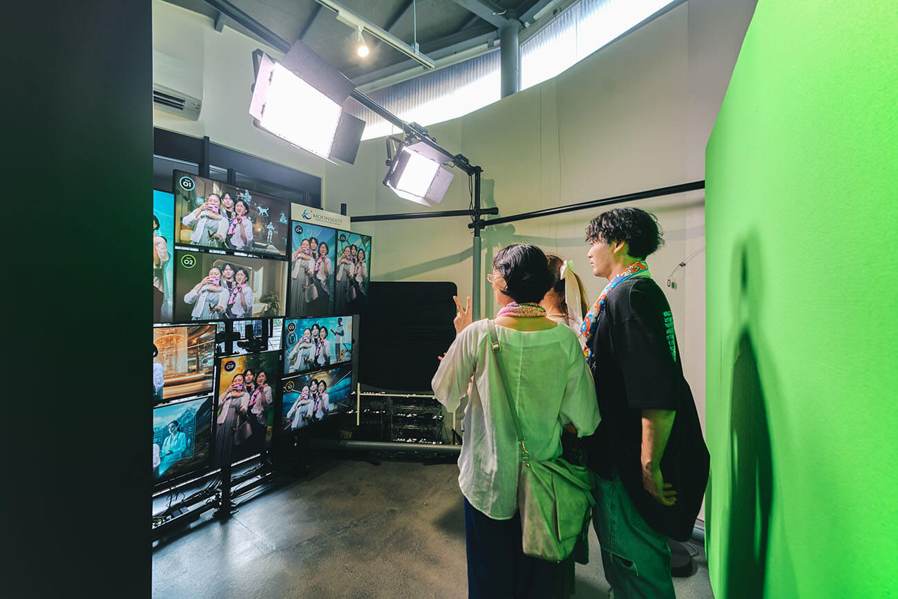 フォトスポットで、実際に参加者が記念撮影を行う様子。男女3名が、万博グッズを身纏いながら、一人がスマートフォンを構えて撮影を行なっている。三人の集合した様子が、10個のディスプレイにそれぞれ表示されている。