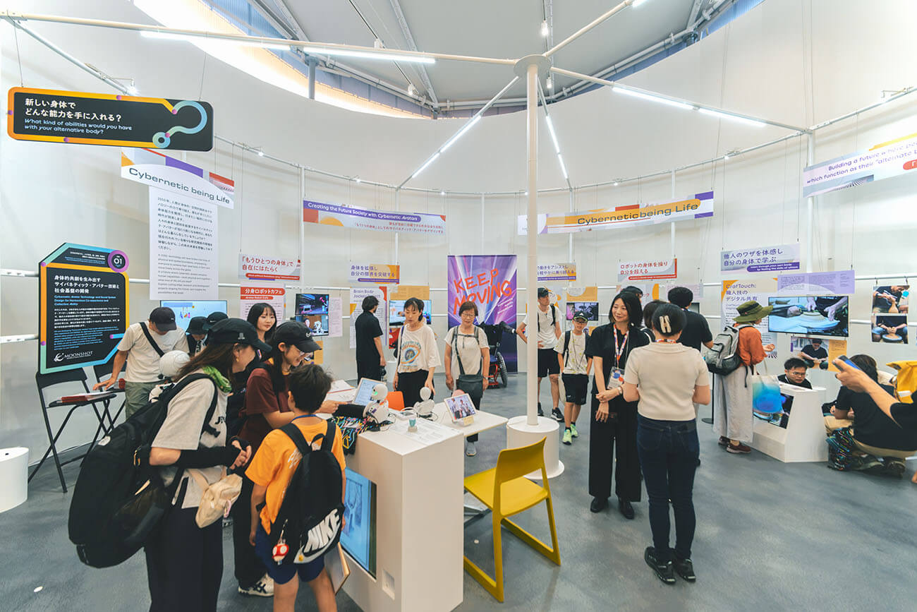 若者や親子連れが体験型展示を巡り、サイバネティック技術のパネルやデモを興味深く見学しているにぎわいある展示空間の様子。