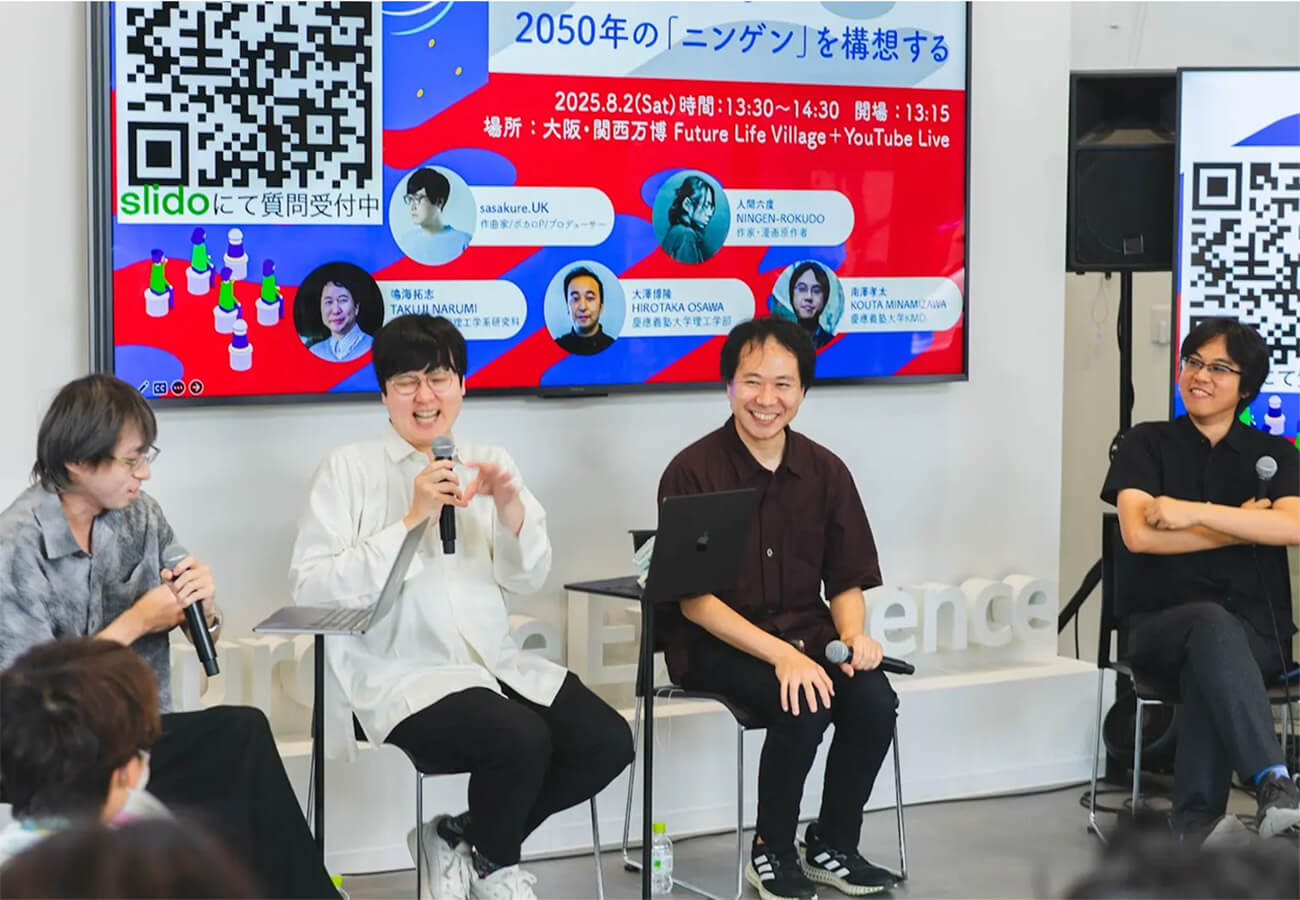 研究者やクリエイターが壇上でマイクを手に議論し、来場者が聞き入るステージイベントの様子。背景スクリーンには「2050年のニンゲンを構想する」企画の案内が表示されている。