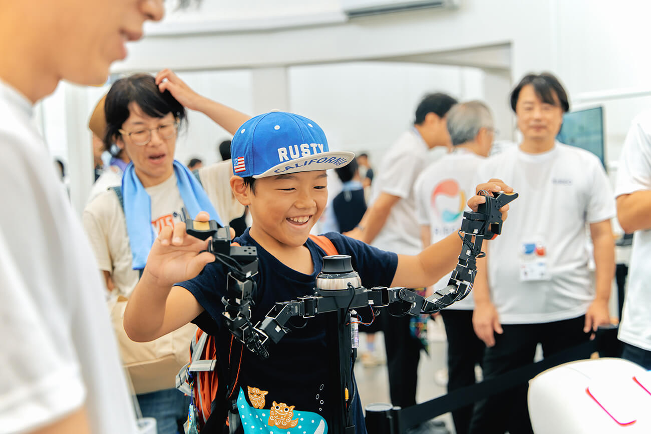 帽子をかぶった男の子が多関節ロボットアームを操作し、笑顔で動作を試している。周囲では大人たちが見守り、来場者が先端技術を体験する展示の一場面。