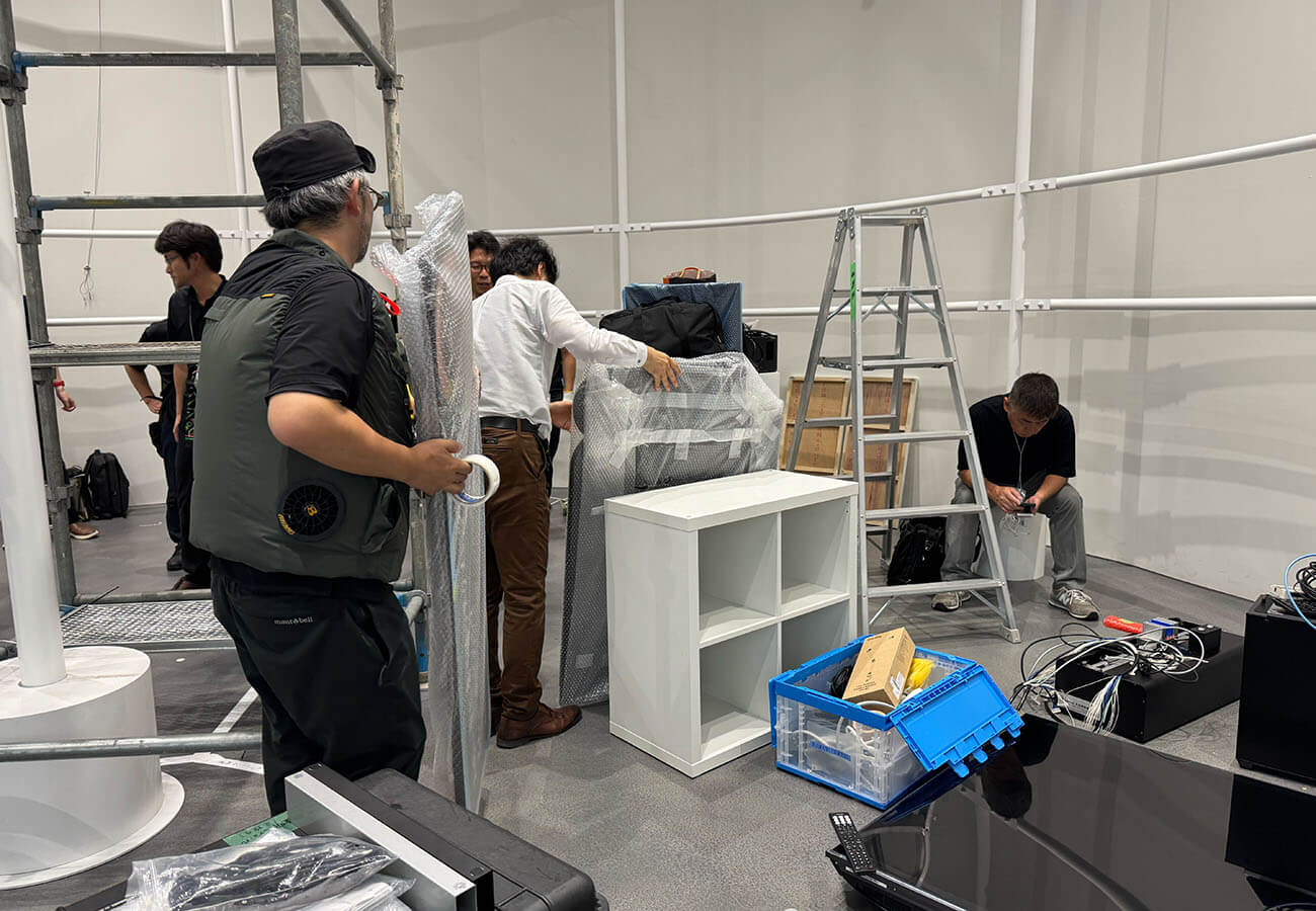 スタッフが棚や機材を梱包し、脚立や資材が並ぶ中で展示の準備を進めている設営作業の様子。週替わり展示の入れ替えを支える裏側を写す。
