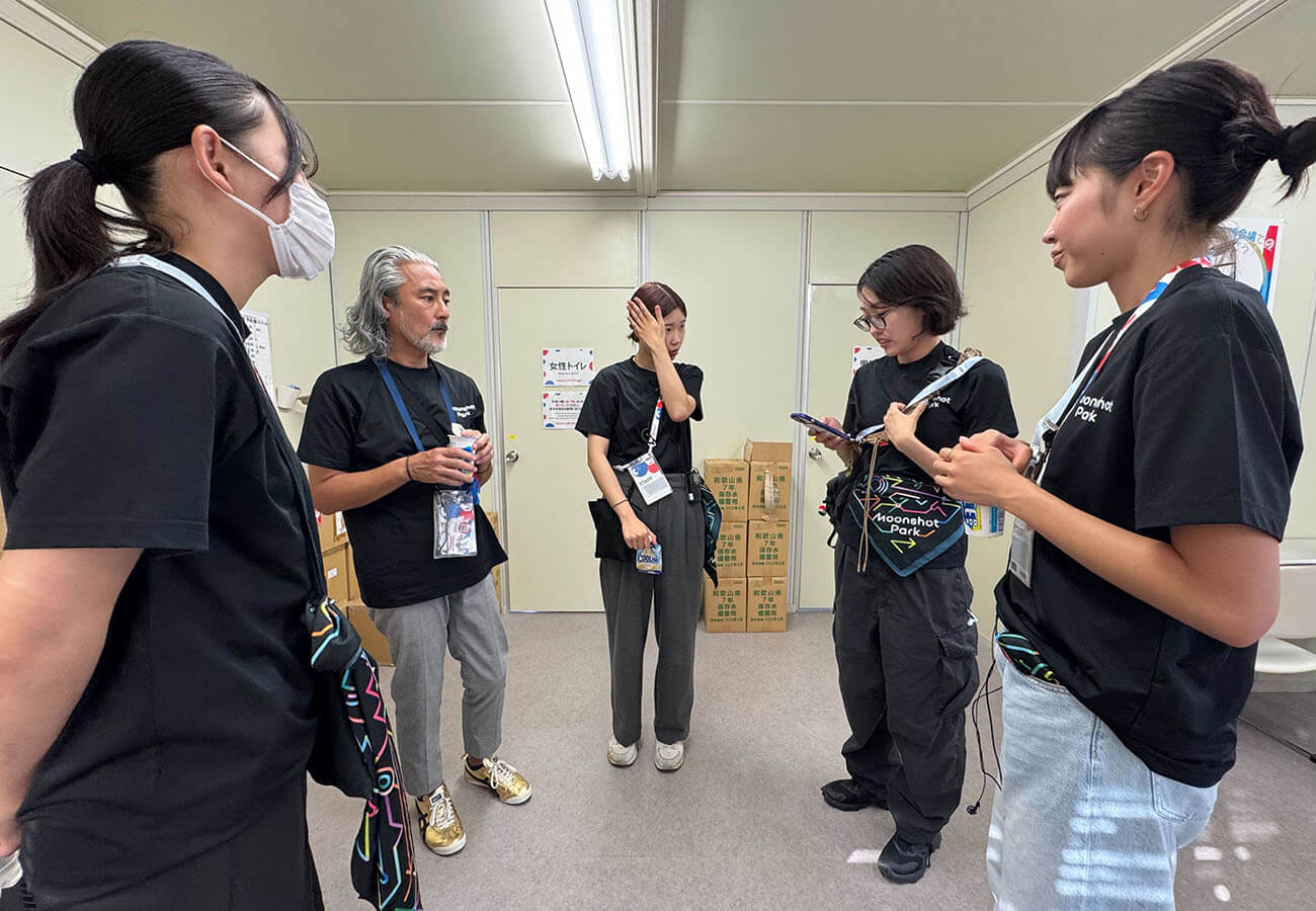 黒いスタッフTシャツを着たメンバーが控室で立ち話をし、担当内容の確認や共有を行っている様子。展示運営前のオリエンテーションの場面。