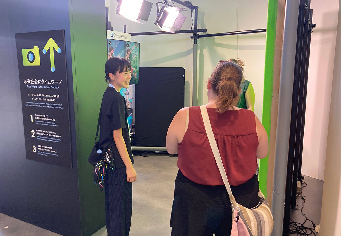 撮影ブースで海外からの来場者がスタッフの案内を受け、未来社会の背景に自分を投影する体験を準備している様子。ライトが照らす中、黒い案内パネルが設置されている。