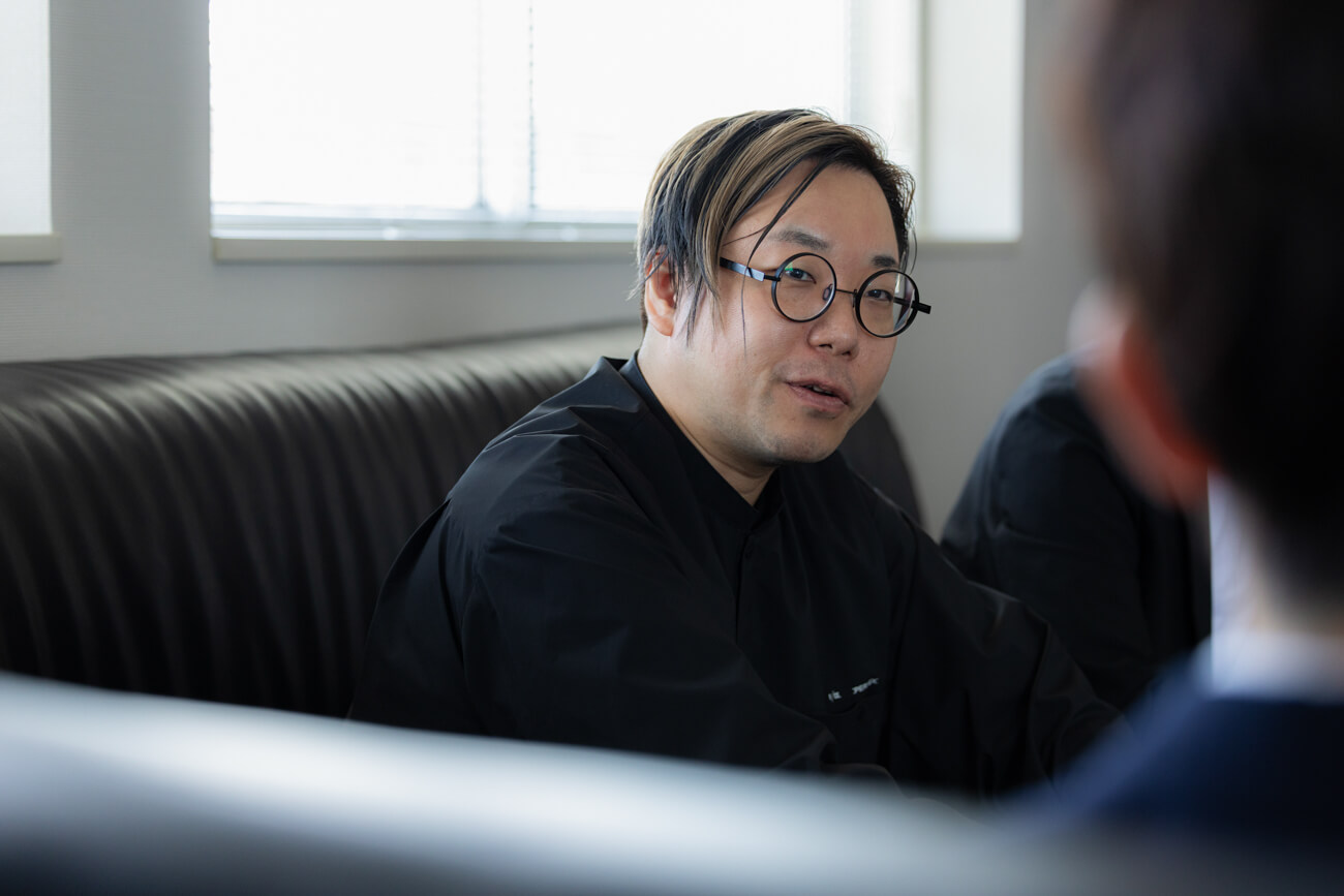 応接室のソファに座る男性が、インタビューイーに対して話しかけている様子