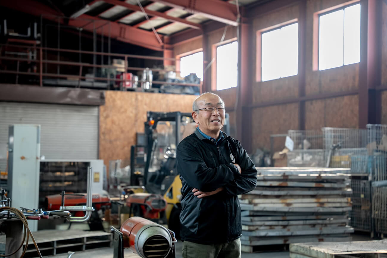 セイカンの元専務・高橋さんが倉庫に立っている様子。笑顔で腕組みをしている