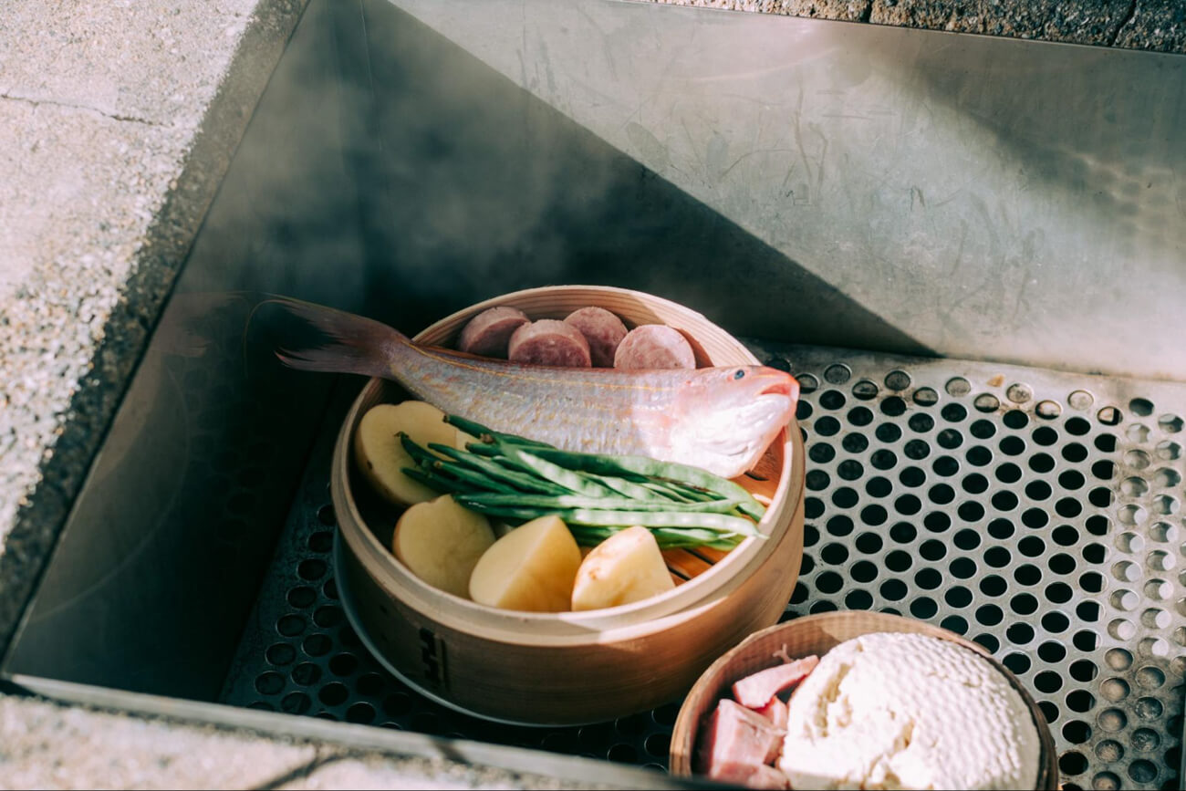 温泉の湯気が湧き上がる網の上に、食材がたっぷり入ったせいろが置かれて、蒸されている写真。美味しそうな鯛が蒸しあげられている