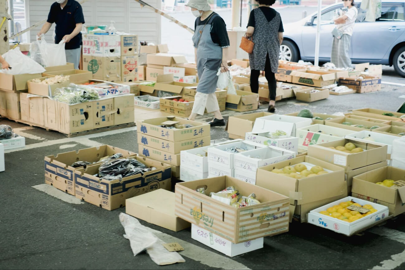 朝市の風景。段ボールいっぱいに入った野菜や果物が地面に並べられ、その間をお客さんが歩いている