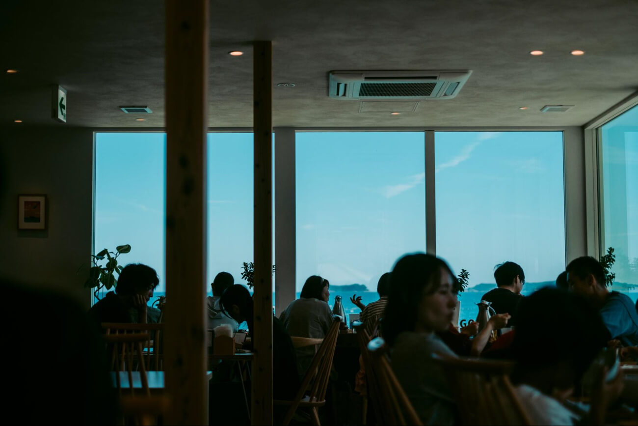 飲食店の店内の写真。食事を楽しむ人々の向こうには、窓越しにあおい海と広い空が見える