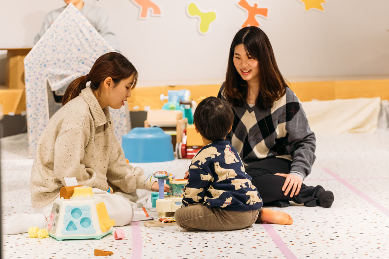 若者世代が、ちいさな赤ん坊と遊んでいる様子。子どもはおもちゃの散らばった床に座って、若者の顔を見つめている
