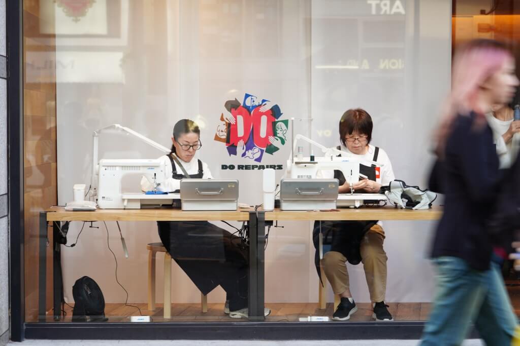 DO REPAIRS参加店舗のシーン。二人のリペアラーの女性が、ショーウインドーの中で洋服のリペア作業を行なっている。
