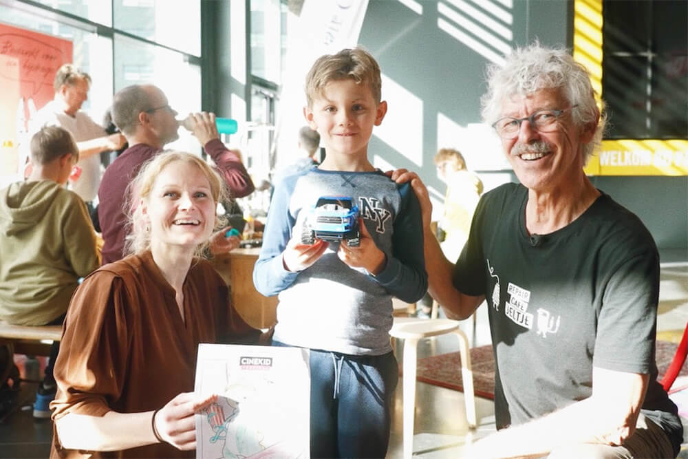 アムステルダムのリペアカフェの写真。男の子が修理してもらったおもちゃを持って微笑んでいる。男の子の右側にリペアラーの白髪の男性が笑顔で座っている。左には男の子の母親が微笑んでいる。