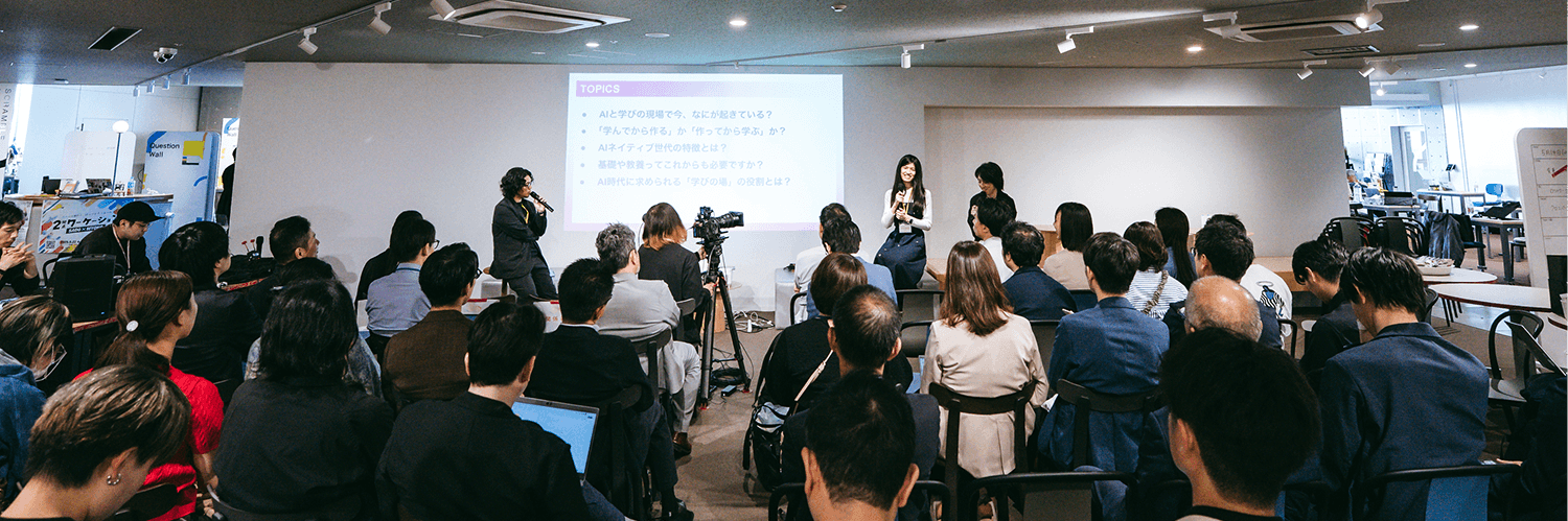 写真：別の会場で開催されたトークセッションの様子。スクリーンを背景に登壇者3名がマイクを持って対話し、50名ほどの参加者が前方に集まって聴講している。