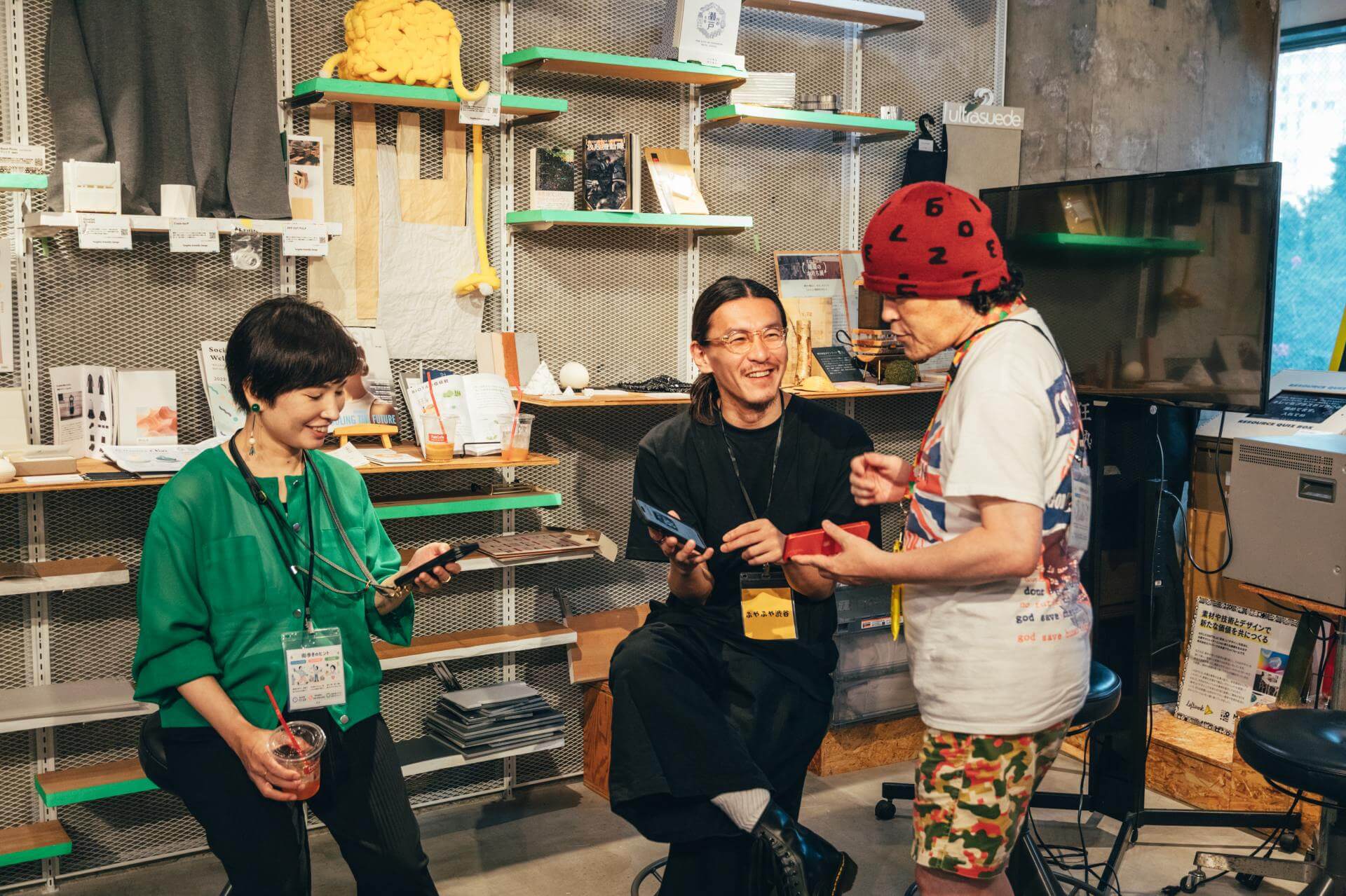 素材見本が並ぶ棚を背に、3人がそれぞれスマートフォンを手に笑いながら話している
