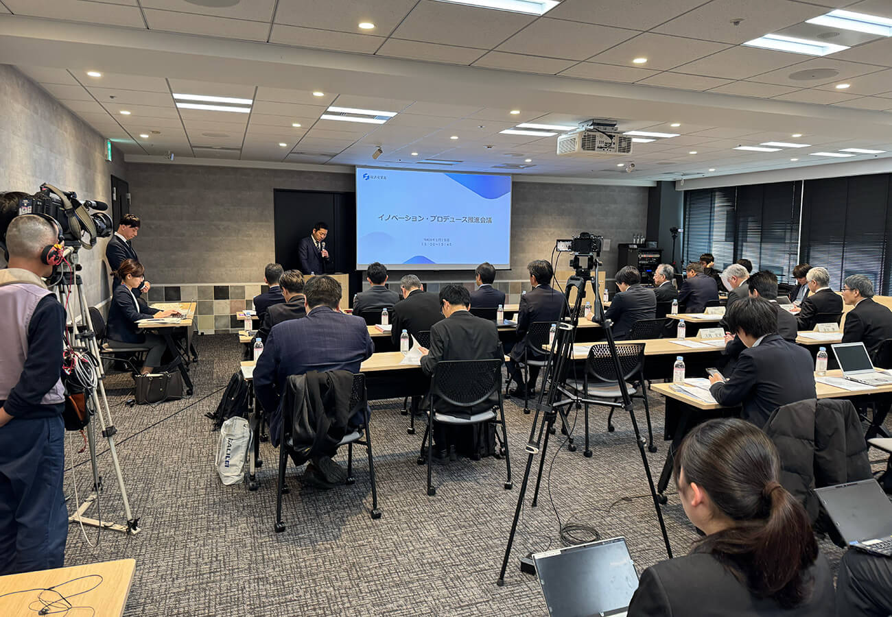 「イノベーション・プロデュース推進会議」の会場。スクリーンに会議名と令和8年1月19日の日付が映され、演台で話す登壇者に向かって多数の出席者が着席している。後方には複数のテレビカメラが並ぶ。