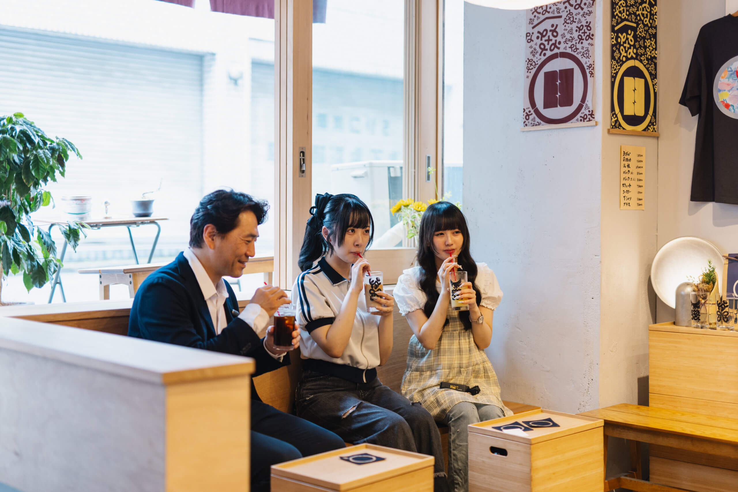 まちあるきの取材風景。明るい採光のカフェのなかで、教授1人と学生2人が並んでベンチに座り、ドリンクを飲んでいる