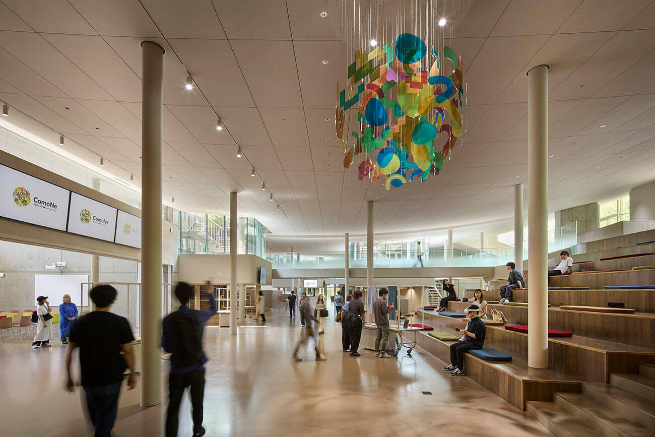 A spacious shared area with a soaring ceiling. People relax on grand staircase-like viewing seats, while a vibrant hanging sculpture adorns the center.