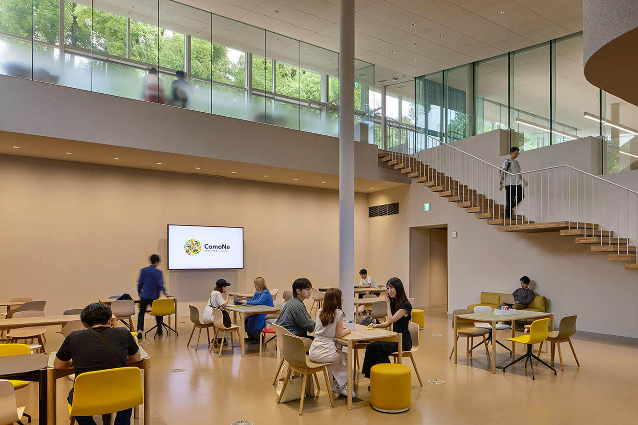 The collaborative space. People are discussing and working at the table seating, and the open space features stairs and large windows.
