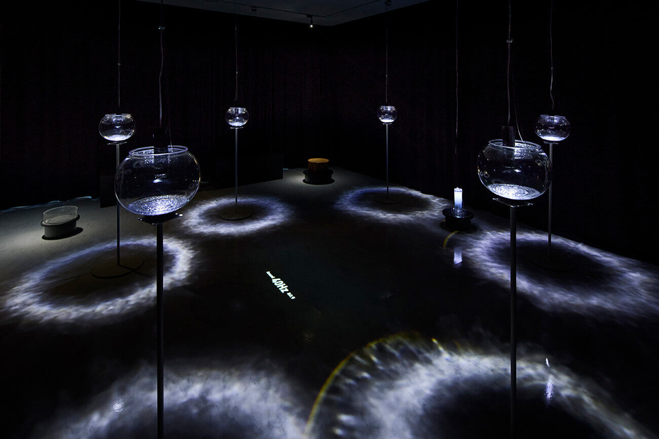 インスタレーション《Deep ground Resonance》の展示風景。吊り下げられたガラス容器と床に広がる光の波紋が広がる作品。