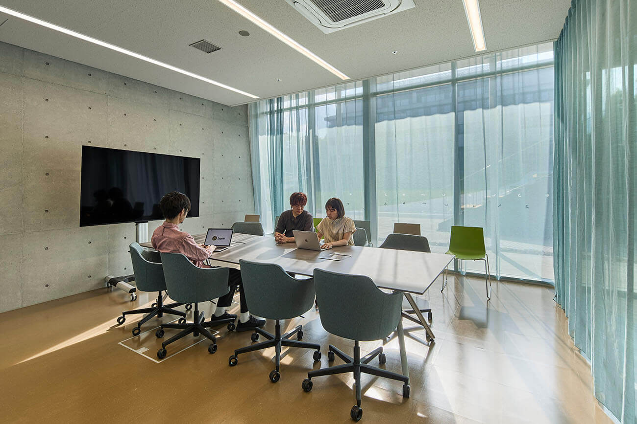 A meeting room bathed in natural light streaming through large windows. Movable tables and casters-equipped chairs are arranged, with several people spread out with their laptops discussing matters.