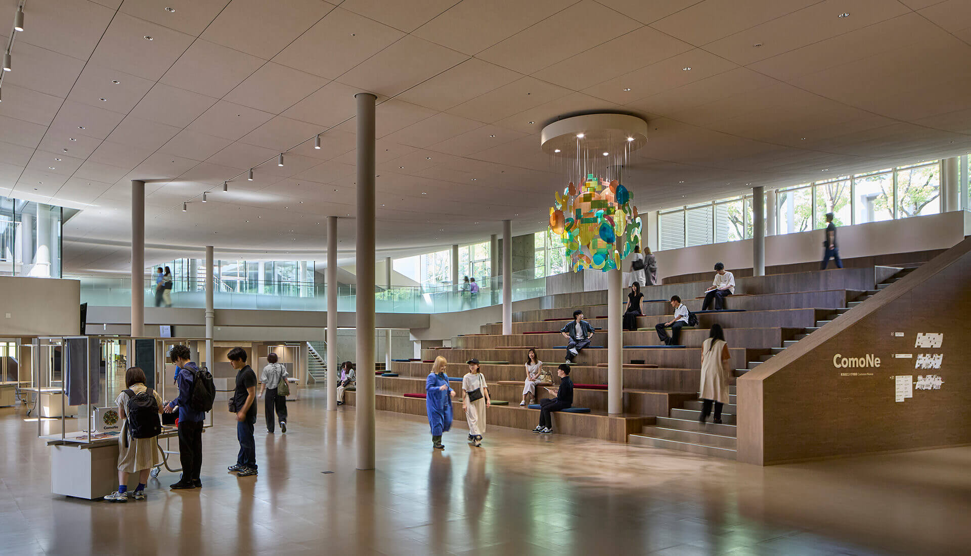 Inside ComoNe. A spacious double-height atrium features grand staircase-like viewing seats where students and visitors relax at their leisure. Colorful art installations hang from the central ceiling.