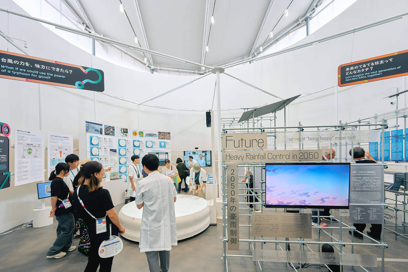 学生や家族連れが「豪雨制御」など気象研究のパネルや映像展示を見ながら歩き回り、スタッフと対話する様子が広がる明るい展示空間。